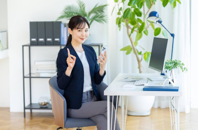 スマホを持って人差し指を立てているオフィスカジュアルの女性